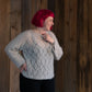 A person with bright red hair smiles and looks to the side, standing in front of a wooden panel wall. They are wearing a vintage-inspired Atmosphere Lace Mohair Pullover by Halcyon Yarn in light gray with a diamond pattern, paired with dark pants. Their hand rests on their hip, showcasing multiple rings on their fingers.