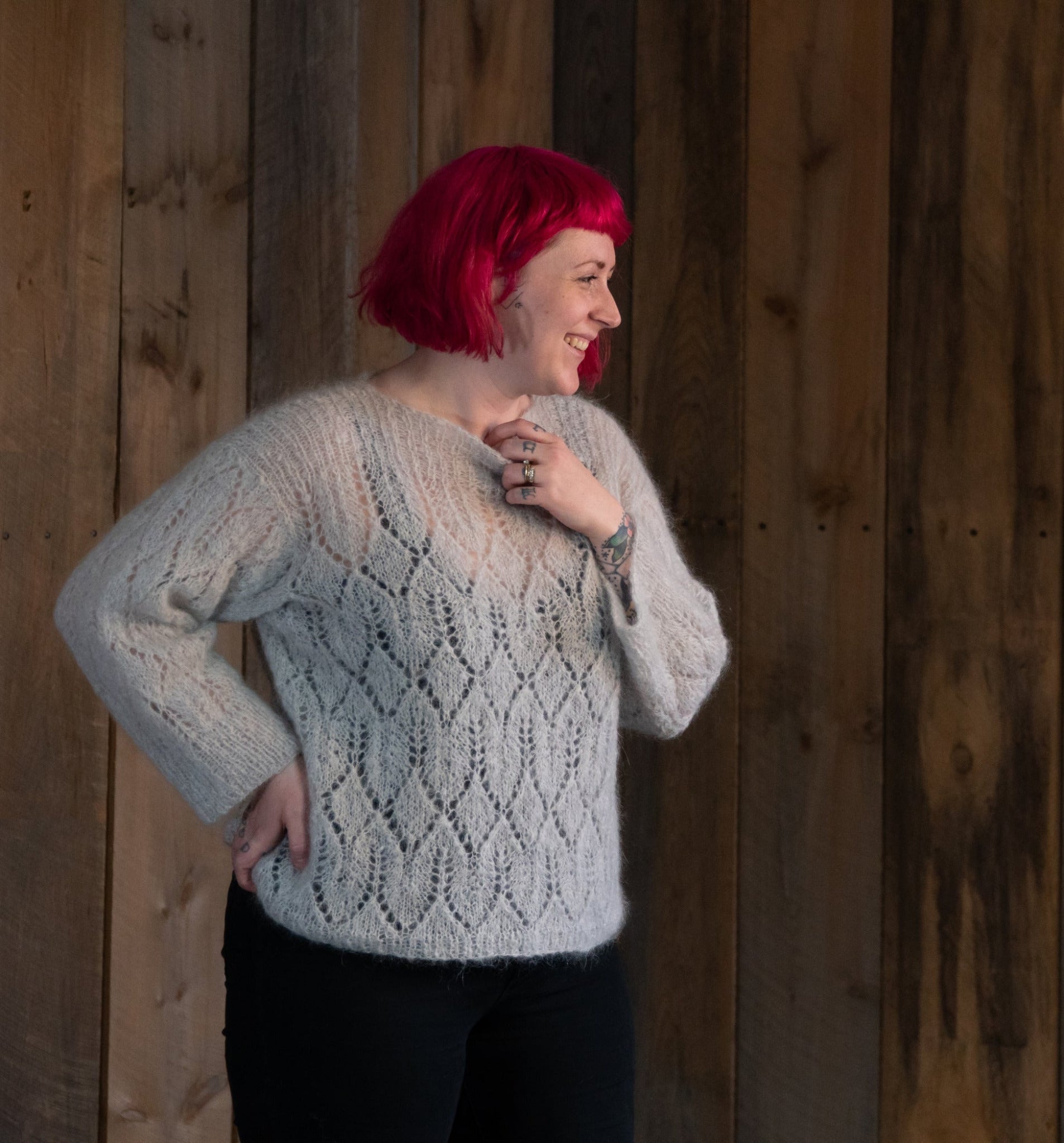 A woman with bright pink hair smiles while wearing the Atmosphere Lace Mohair Pullover from Halcyon Yarn. She is standing in front of a wooden wall with her right hand on her hip, showcasing several rings on her fingers and a visible tattoo on her left forearm.