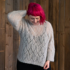 A person with bright pink hair is smiling while looking down and touching the back of their neck with one hand. They are wearing a cozy, light-colored, vintage-inspired Atmosphere Lace Mohair Pullover from Halcyon Yarn and standing in front of a wooden wall.