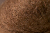 Close-up of a brown, fibrous texture, resembling the surface of densely packed natural fibers, such as Kingfisher Yarn & Fibre’s Jo Sharp Rare Comfort Kid Mohair. The fibers vary slightly in color, giving a textured and somewhat uneven appearance that evokes rare comfort found in airy sweaters.