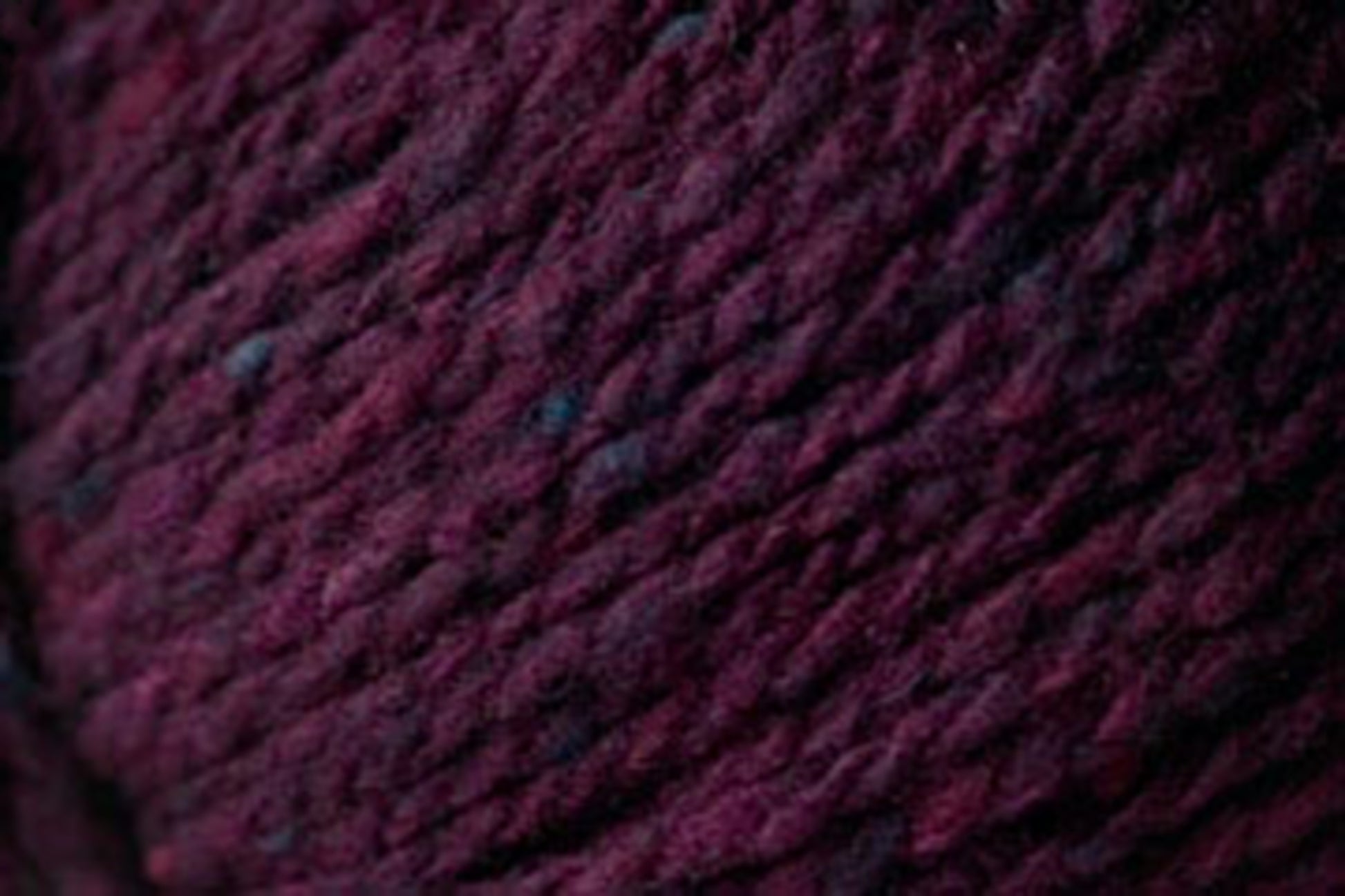 Close-up image of thick, dark burgundy Kingfisher Yarn & Fibre's Jo Sharp Silkroad Aran Tweed with subtle variations in color, displaying a textured, woven appearance. The yarn's fibers are slightly mottled, indicating a rich and soft texture that is suitable for knitting or crocheting.