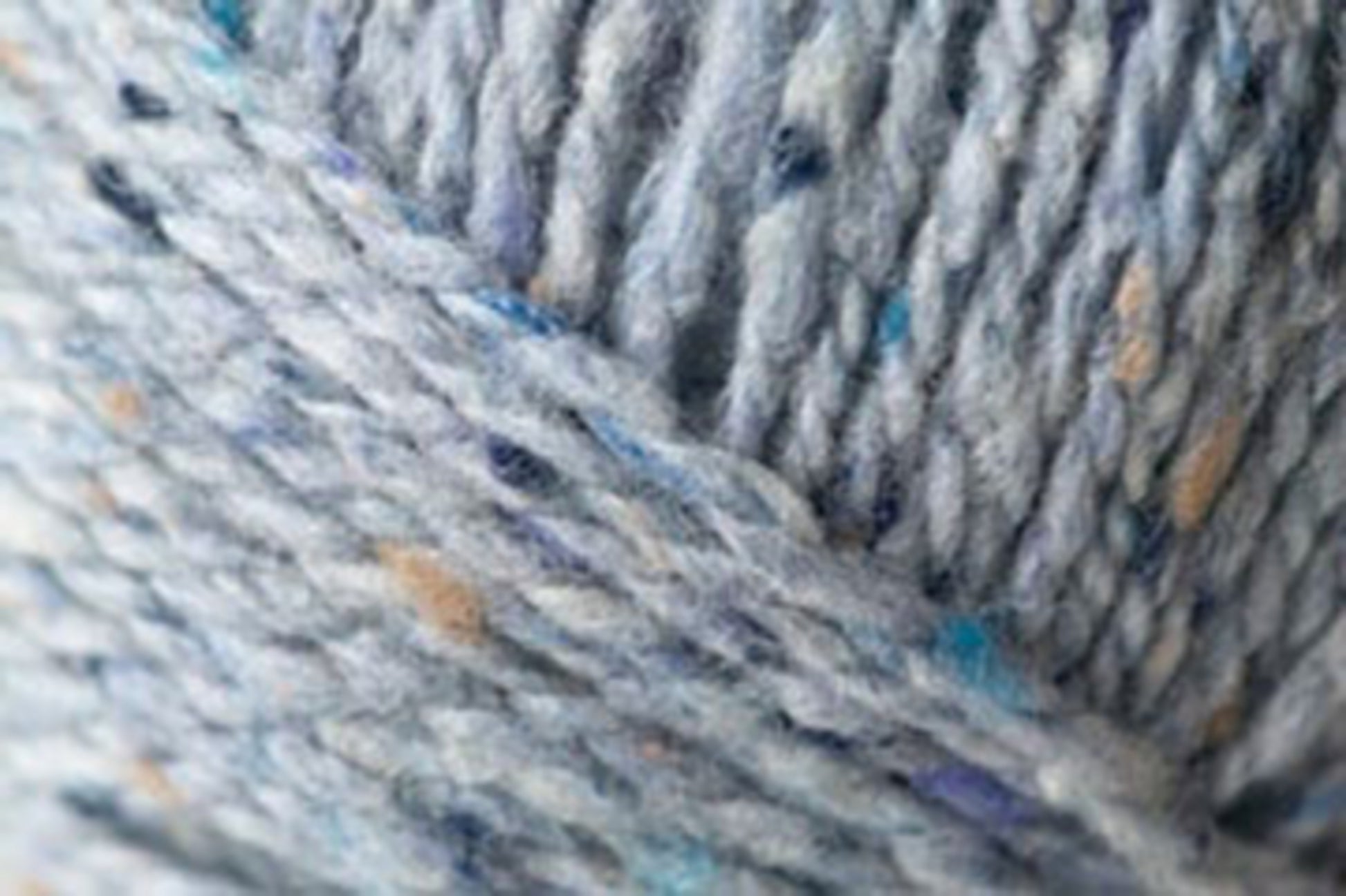 Close-up of the luxurious Kingfisher Yarn & Fibre Jo Sharp Silkroad Aran Tweed yarn featuring specks of blue, beige, and white. The image showcases tightly woven strands forming a textured pattern. The yarn appears soft and slightly fuzzy, making it ideal for knitting or crocheting projects.