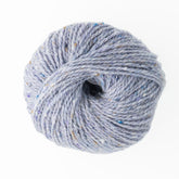 A ball of Jo Sharp Silkroad Aran Tweed yarn in light gray, speckled with blue, brown, and beige, is showcased against a white background. This luxurious product from Kingfisher Yarn & Fibre features a tightly spun blend of wool, silk, and cashmere, forming a neat and compact ball.