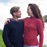 A man and a woman stand outdoors near the water, both wearing beautifully knitted sweaters from Twist Collective's Mosey Cabled Pullover collection. The man wears a dark sweater with saddle shoulders, while the woman dons a red one that showcases an intricate cable pattern. They are smiling at each other, with the woman's arm around his shoulders. Green grass and trees are in the background.