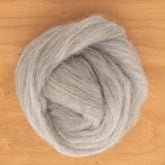 A coiled bundle of Shetland Top Wool Fiber from Europa Wools Ltd lies on a wooden surface. The soft, gray fibers appear light, airy, and fluffy, with a smooth, slightly fuzzy texture. The natural gray color contrasts gently with the warm tones of the wood background, making it perfect for delicate lace knitting projects.