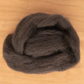 A coil of Dashing Mouse's Black Welsh Top Wool Fiber, perfect for spinning yarn, is placed on a light wood surface. The wool has a fluffy and textured appearance, loosely arranged in a circular shape.