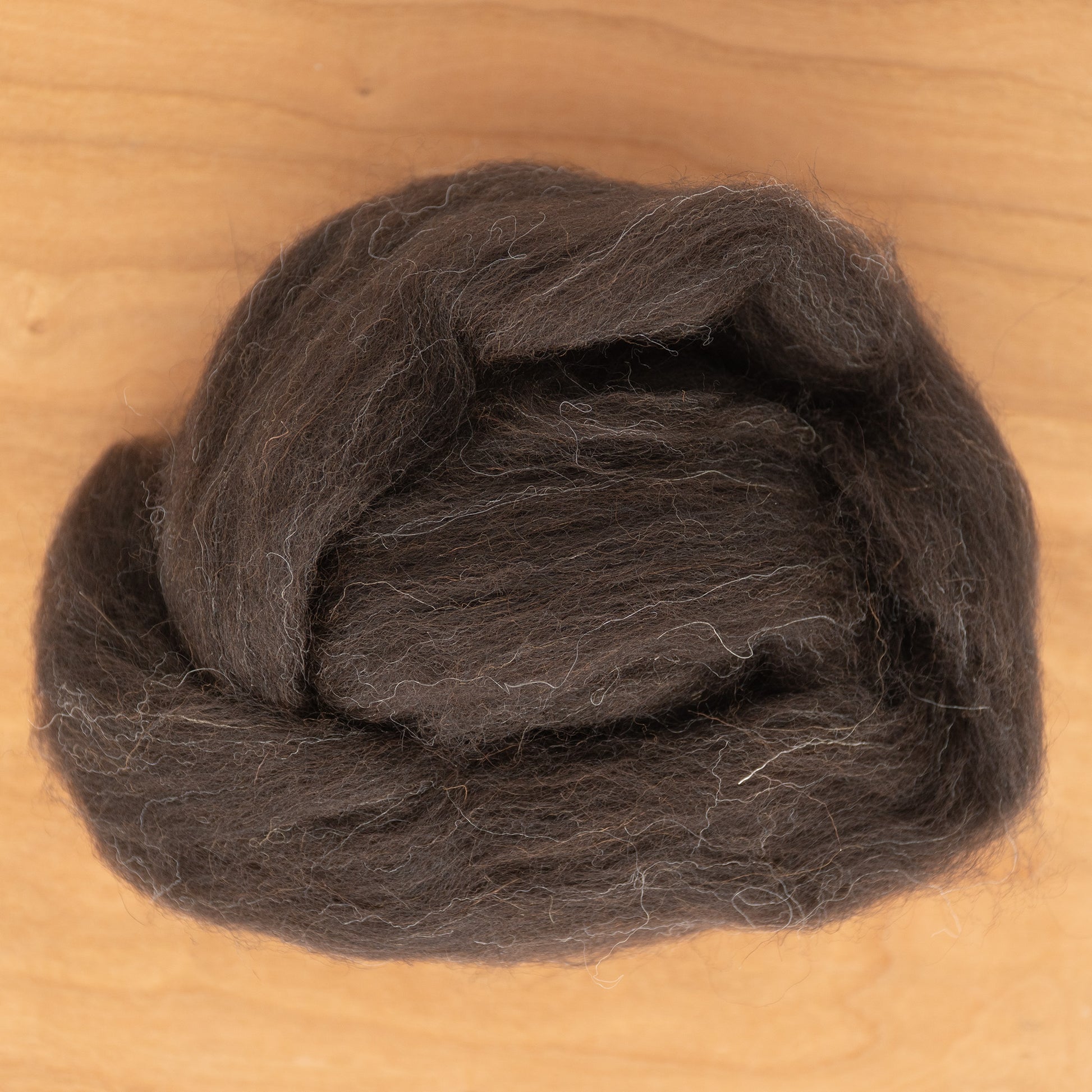 A coil of Dashing Mouse's Black Welsh Top Wool Fiber lies neatly on a light wooden surface. The dark brown wool roving has a soft, slightly fuzzy texture, with a few stray fibers visible.