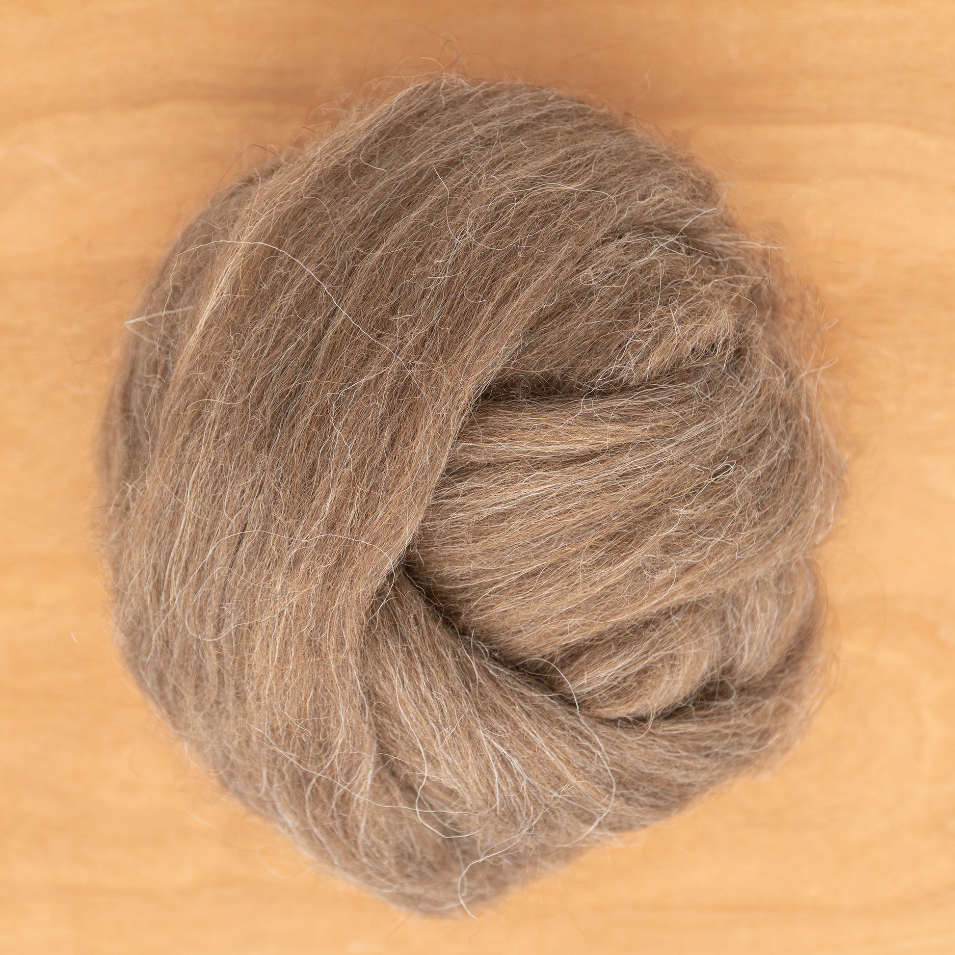 A ball of Icelandic Top Wool Fiber from Europa Wools Ltd, sold by the ounce, is loosely coiled on a wooden surface. Its soft and fluffy texture showcases the charm of dual-coated fleece with visible mixes of light and dark brown tog fibers.