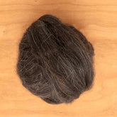 A round bundle of Icelandic Top Wool Fiber, characterized by dark brown tones with gray strands from an Icelandic sheep's dual-coated fleece, rests on a wooden surface. This fiber is offered by Europa Wools Ltd and sold by the ounce.