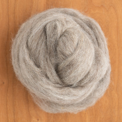 A close-up of a bundle of Coopworth Blend Wool Roving by Dashing Mouse, featuring soft, light gray fibers from New Zealand breeds, placed on a wooden surface. The roving is neatly coiled, showcasing a fluffy texture ideal for spinning or crafting.