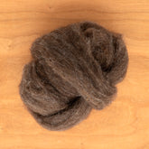 A ball of Dashing Mouse Coopworth Blend Wool Roving rests on a wooden surface. The fibers are loosely gathered, showcasing light and dark brown tones from New Zealand breeds, creating a textured and soft appearance, perfect for spinning enthusiasts.