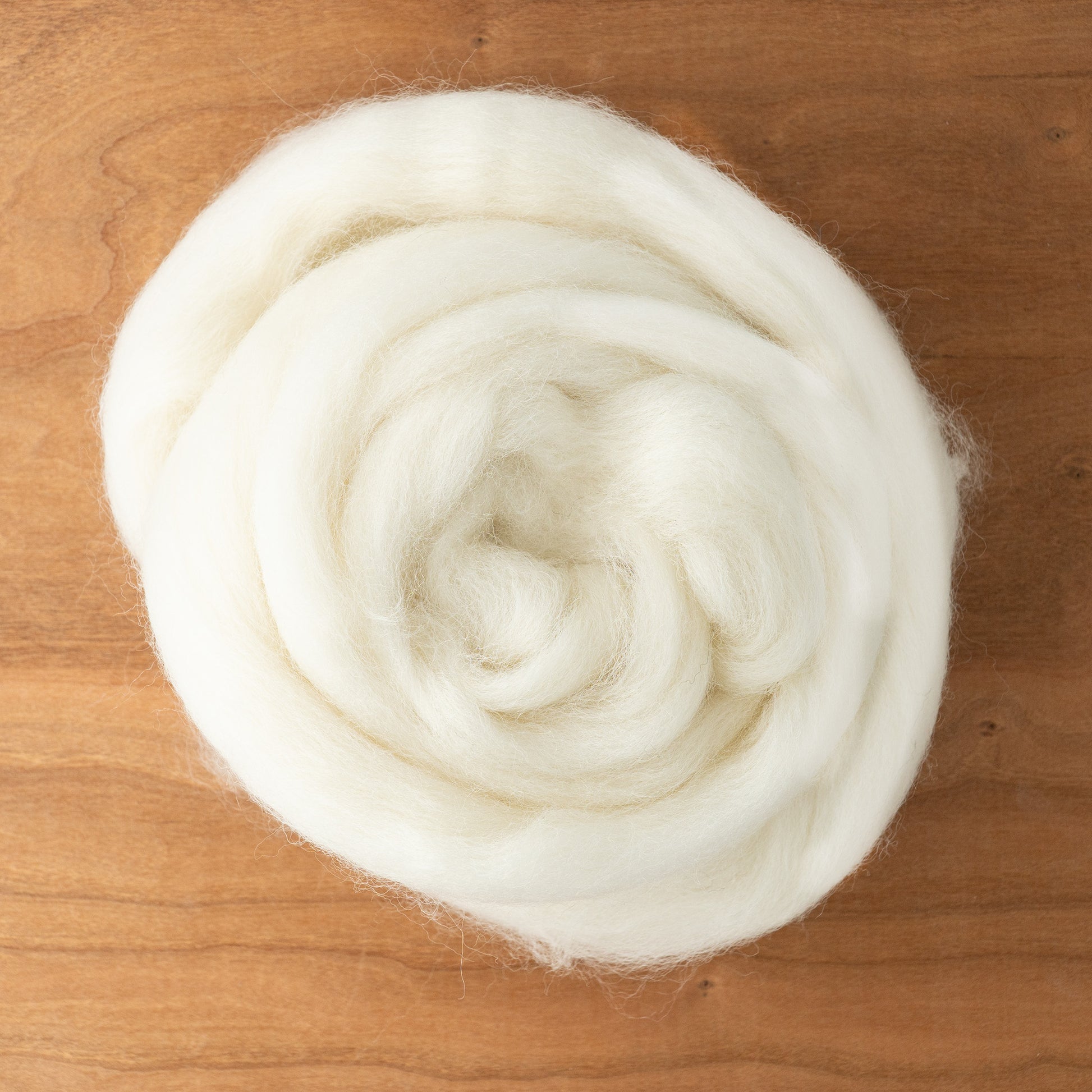 A close-up of a neatly rolled bundle of soft, white RH Lindsay Corriedale Wool Top on a wooden surface. Ideal for needle felting, this wool has a fluffy texture and a slightly spiral shape.