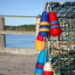 Halcyon Yarn's Lobster Buoy Felted Ornaments hang on a metal trap placed on a wooden dock, set against the backdrop of a calm body of water and a blurred shoreline with green trees under a clear blue sky, evoking the charm of coastal Maine.