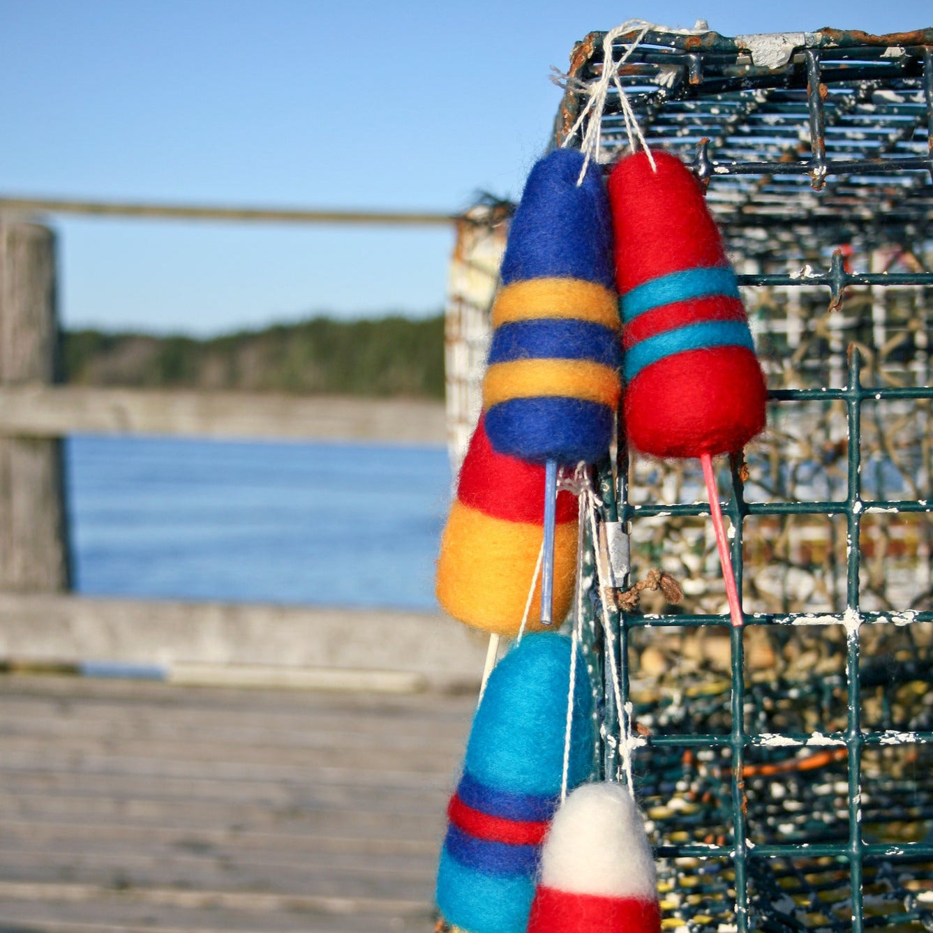 A close-up of brightly colored Lobster Buoy Felted Ornaments by Halcyon Yarn, attached to a wire cage on a wooden dock. Reminiscent of Christmas ornaments, the red, blue, yellow, and white buoys add charm to the coastal Maine scene. The dock overlooks a calm body of water with trees visible in the background under a clear blue sky.