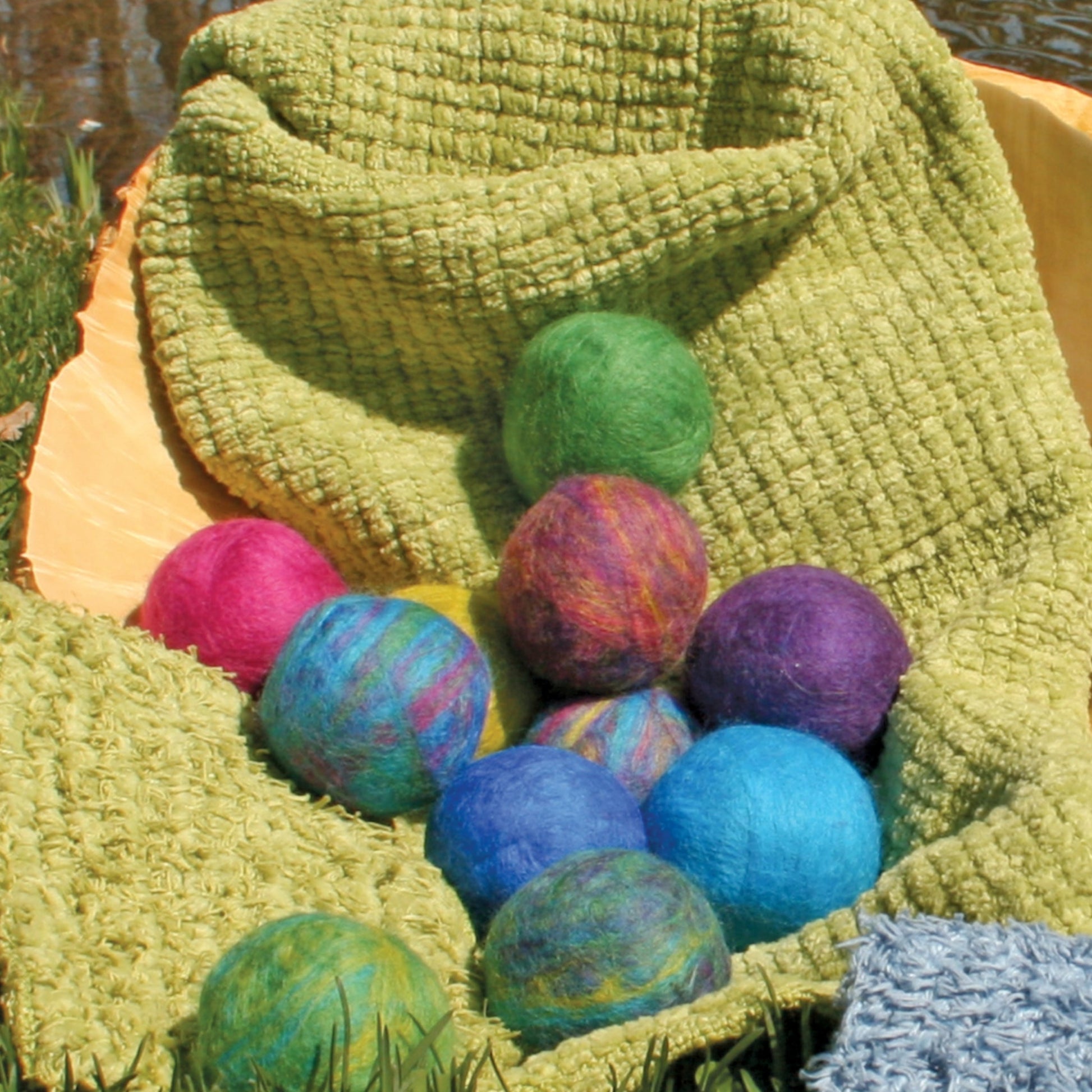 Halcyon Yarn's Felted Soap Balls, showcasing an array of colors including shades of pink, purple, blue, green, and vibrant multicolored patterns, are elegantly arranged on a textured green blanket atop bright yellow fabric in a scenic outdoor grassy setting. Twigs and grass enhance the foreground, creating an inviting scene perfect for all ages.