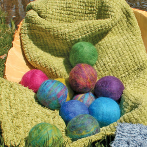 Halcyon Yarn's felted soap balls in shades of green, blue, purple, pink, and multicolor are displayed on a textured green blanket set on a grassy surface. An orange cloth is partially visible in the background. These vibrant creations transform ordinary soap into something extraordinary and are suitable for all ages.