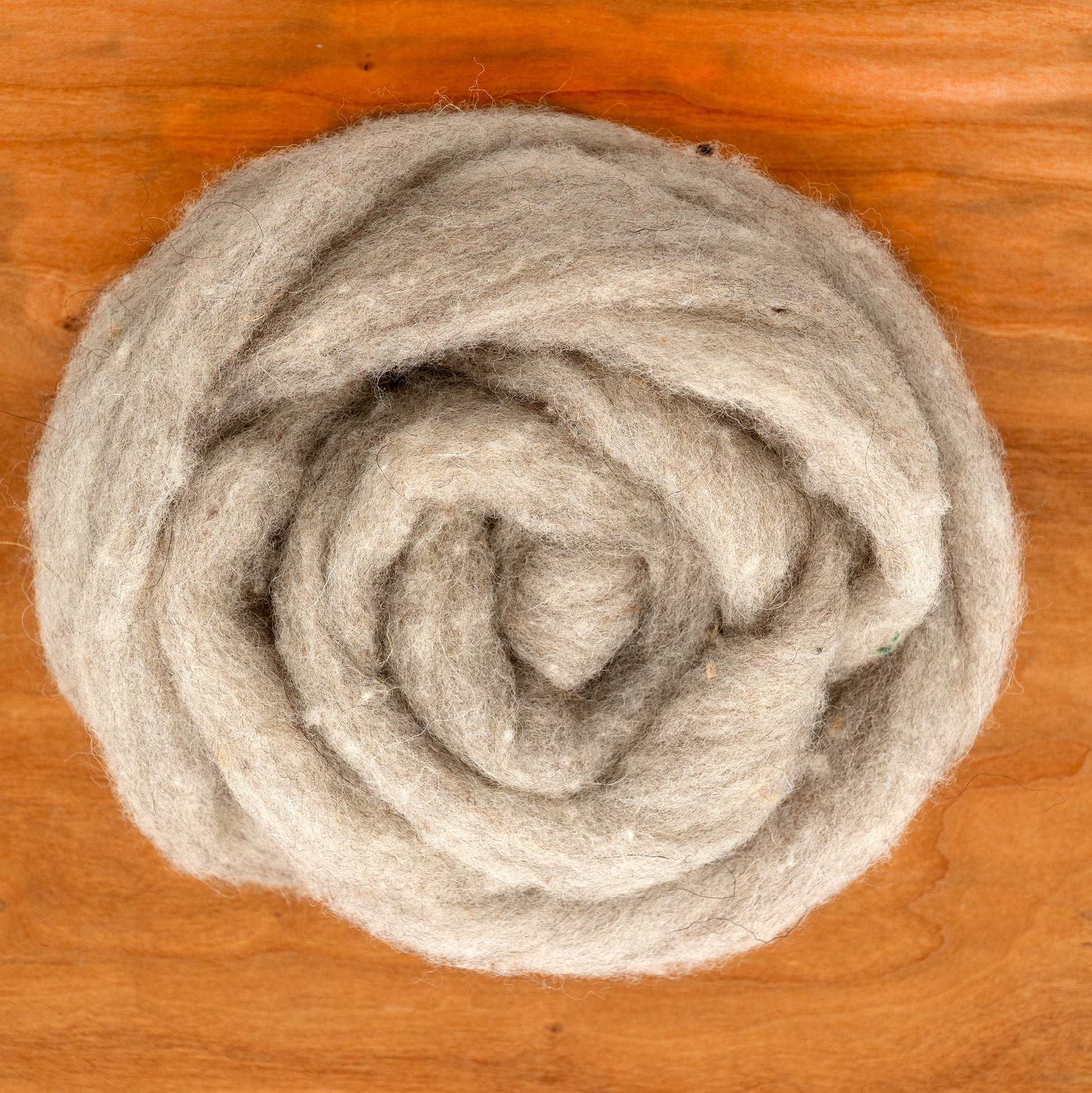 A close-up of RH Lindsay's Romney Wool Fiber Top in a Grey Blend, coiled into a soft spiral on a wooden surface. Ideal for needle felting, this fibrous wool appears fluffy and tightly spiraled in a circular shape.