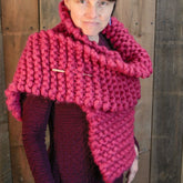 A person wearing The Bold Beauty Wrap, an oversized knit wrap in a vibrant magenta color from Halcyon Yarn. The scarf, made of luxurious unspun wool, is fastened with a shawl pin and draped elegantly over their shoulders. The individual is dressed in a textured garment, standing against a wooden background.