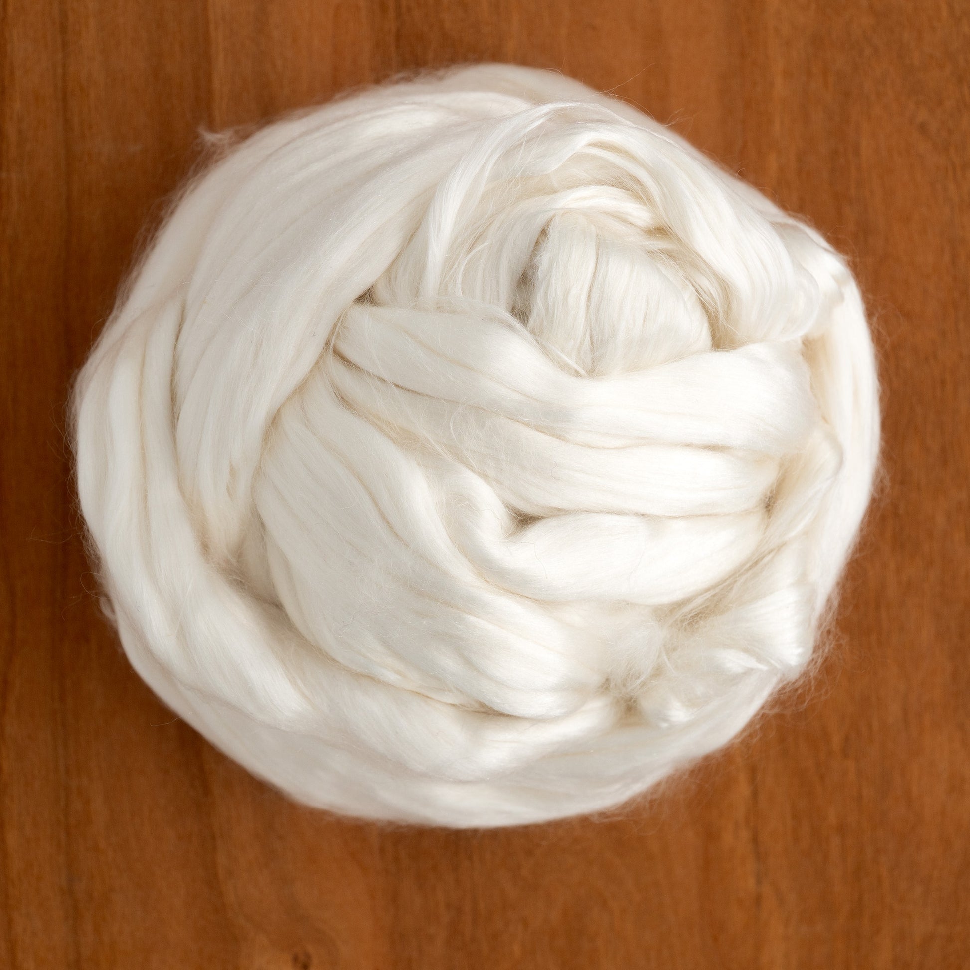 A close-up image of a ball of Europa Wools Ltd's Cultivated Mulberry Silk Top sitting on a wooden surface. The silk is neatly coiled into a soft, round shape, resembling the luxurious texture of 100% silk yarn and highlighting its fibrous, fluffy appearance.