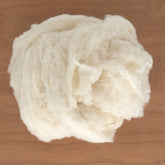 A fluffy, round clump of Europa Wools Ltd's Silk Noil Fiber, Raw Silk (sold by the ounce) is placed on a smooth wooden surface. Much like wool, its slightly fibrous texture and loosely gathered form enhance its natural, airy appearance.