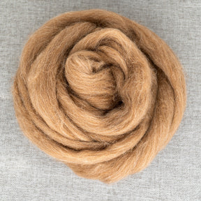 A coiled bundle of light brown Shetland Top Wool Fiber from Europa Wools Ltd is arranged in a circle on a textured gray fabric background. The wool's soft and fluffy texture is clearly visible, creating a warm and cozy appearance that showcases its natural colors.