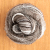 A close-up view of a rolled bundle of Jacob Fiber from Europa Wools Ltd, featuring light and dark gray tones that resemble a soft swirl. This wool, sold by the ounce, is renowned for its varied staple lengths and is placed on a light wooden surface to accentuate its fluffy texture and contrasting shades.