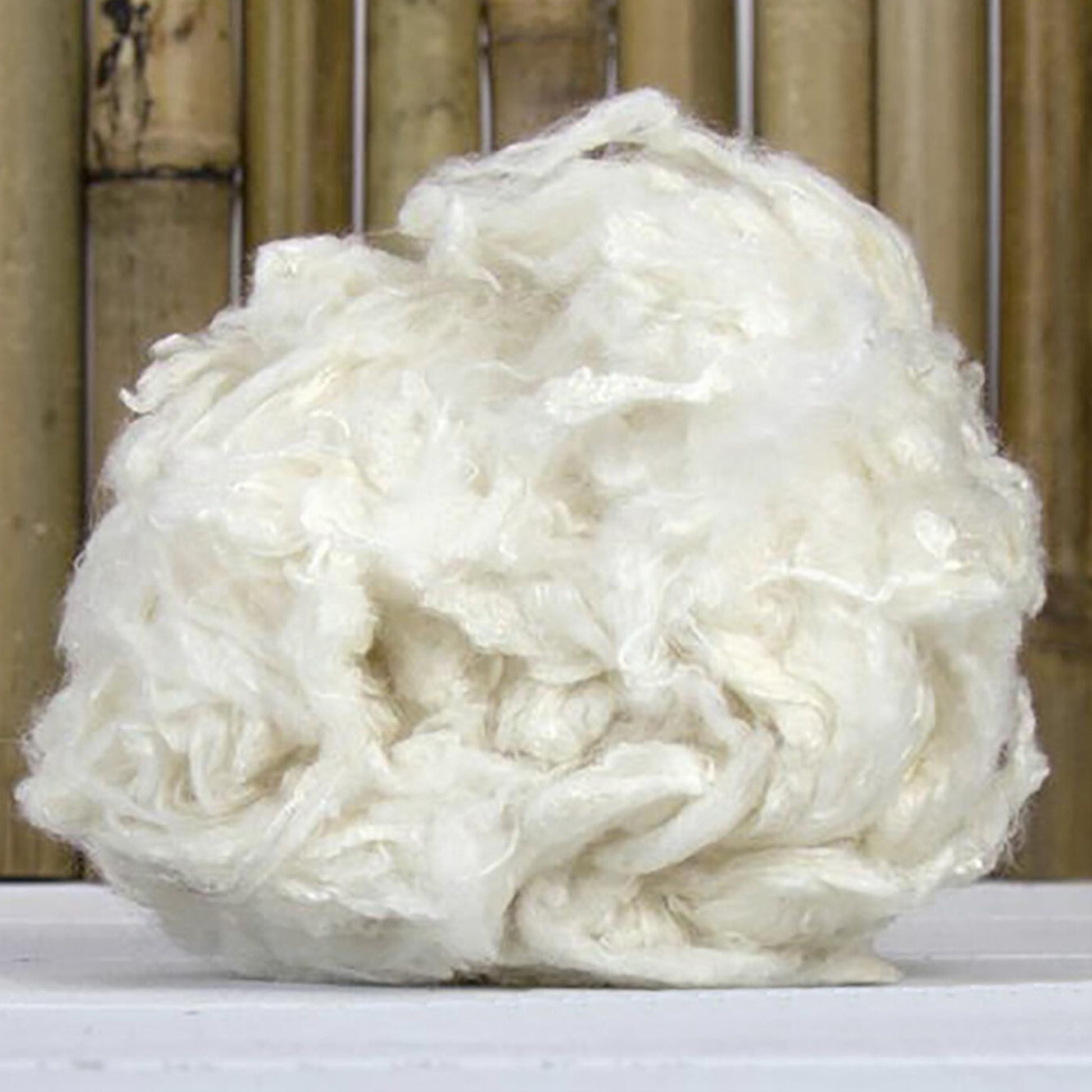 A loosely packed ball of Europa Wools Ltd's Bamboo Fiber sits on a light-colored surface with a background of vertical bamboo stalks. The fiber threads, appearing soft and fluffy with a slightly tangled texture, evoke the natural elegance often found in designer yarns made from sustainable materials.