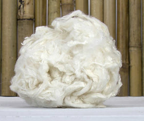 A close-up of a large ball of white Bamboo Fiber from Europa Wools Ltd, resembling cotton or wool, placed on a white surface. The background features vertically arranged bamboo poles. The fibers, an excellent renewable resource, appear soft and fluffy with a slightly tangled texture.