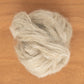 On a wooden surface lies an ounce of Europa Wools Ltd's Flax Roving, with strands of soft, light gray wool fiber exhibiting a fluffy texture. These fibers intertwine loosely, forming an irregular shape reminiscent of a skein.