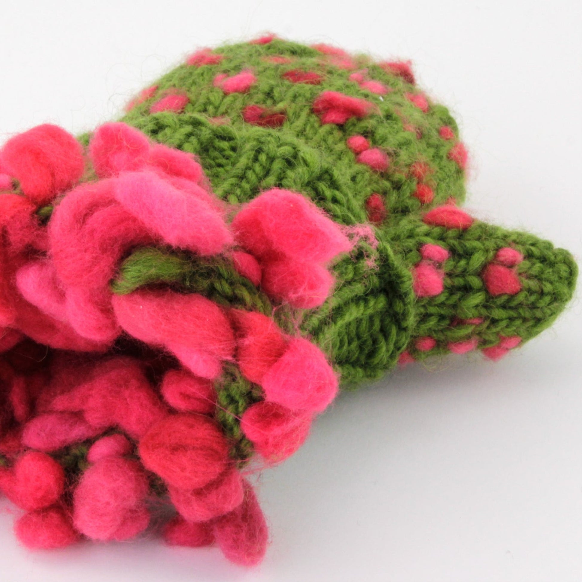 A crocheted item resembling a cactus with vibrant pink flowers, perfect as winter gifts, features a textured green body with pink polka dots against a plain white background. The fluffy, soft yarn adds volume to the flowers, reminiscent of Halcyon Yarn's Snuggly Stuffed Mittens' cozy fleece mittens. Download now!