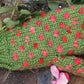 A cozy Snuggly Stuffed Mitten from Halcyon Yarn, designed in green with pink spots to mimic the appearance of a cactus, is lying on a wooden surface. Green thorny branches and a few pink flower petals are scattered around it.