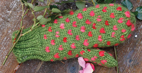 A pair of Snuggly Stuffed Mittens, adorned with pink polka dots, is placed on a wooden surface, partially covered by green leaves and pink rose petals—a charming winter gift suggestion from Halcyon Yarn.