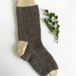 A single sock from the "Classic Socks, download" collection by Yankee Knitter Designs features a gray body with cream-colored toe, heel, and cuff. It lies flat on a white surface, revealing its intricate pattern alongside a small sprig of green leaves.