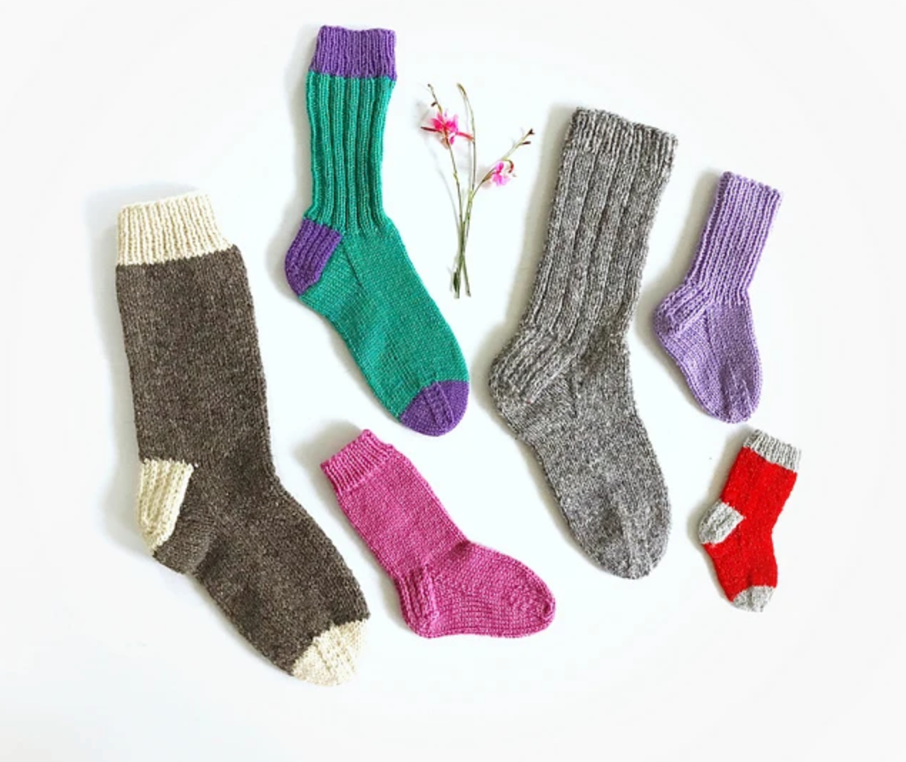 A delightful circle is formed by a collection of Classic Socks from Yankee Knitter Designs, available in an array of sizes and vibrant colors such as gray, green, purple, pink, and red. These socks showcase diverse patterns and various yarn weights. At the center on a pristine white background are two elegantly placed small pink flowers.