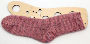 The Classic Socks by Yankee Knitter Designs are showcased on a wooden sock blocker to maintain their shape. These hand-knitted, ribbed socks come in a delightful blend of various shades of pink, creating a beautiful marled effect. The versatile pattern is available for download and can be made in sizes from baby to adult using different yarn weights.