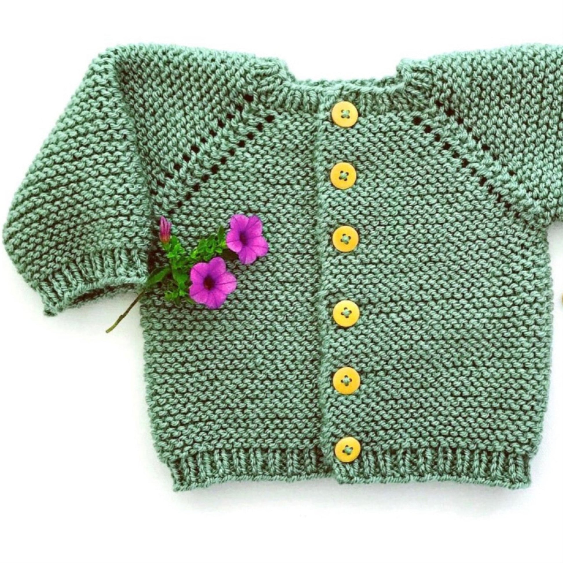 The Etta Cardigan from Yankee Knitter Designs is a hand-knitted baby garment in green, crafted from worsted weight yarn and adorned with yellow buttons. Displayed against a plain background, two purple flowers are tucked into the left sleeve, adding a touch of color and charm. The cardigan features a simple, textured knitting pattern.