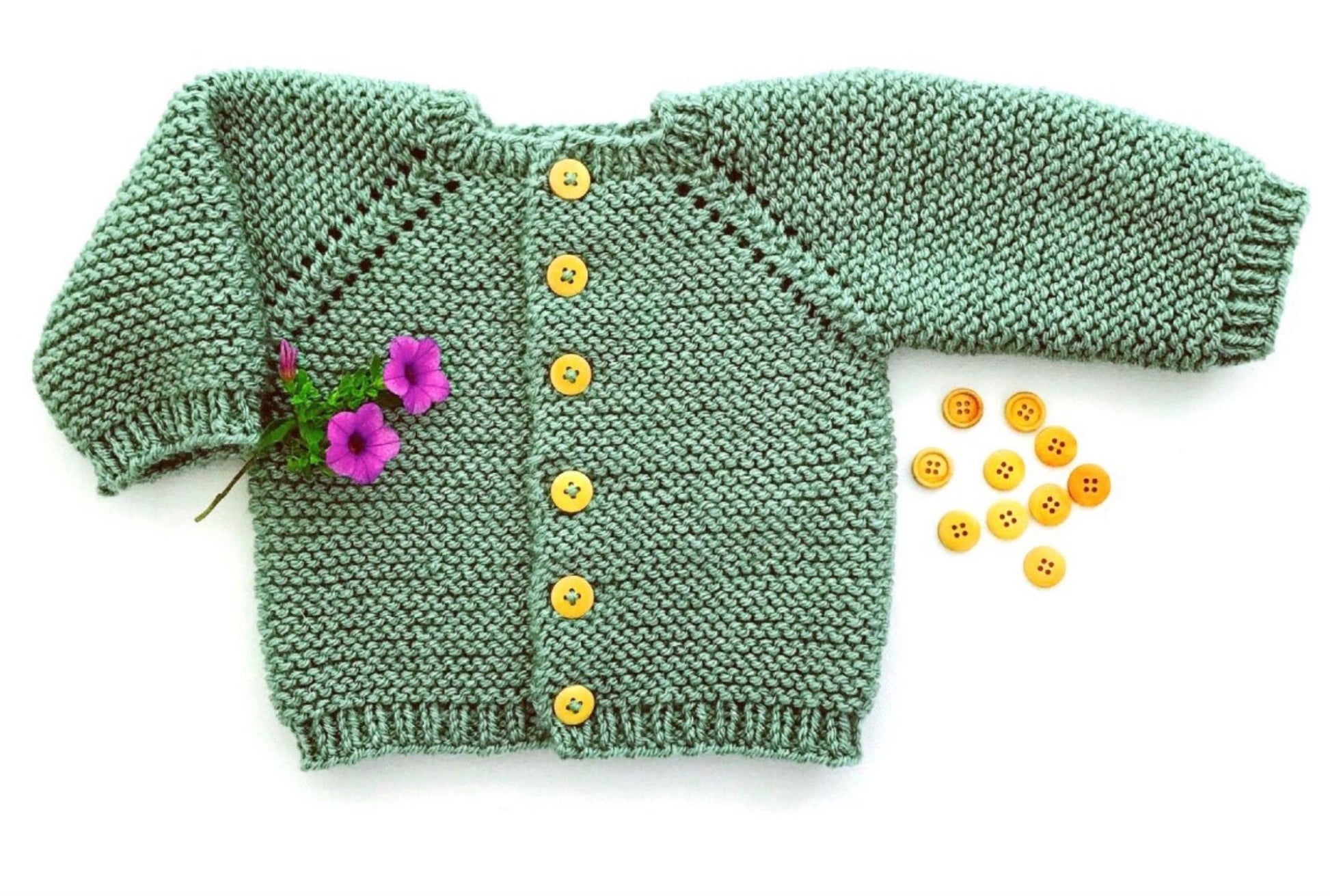 The Etta Cardigan by Yankee Knitter Designs, a hand-knitted green baby cardigan with yellow buttons, constructed from worsted weight yarn, is showcased against a white background. Two purple flowers adorn the left sleeve of the sweater, and several yellow buttons are positioned beside it.