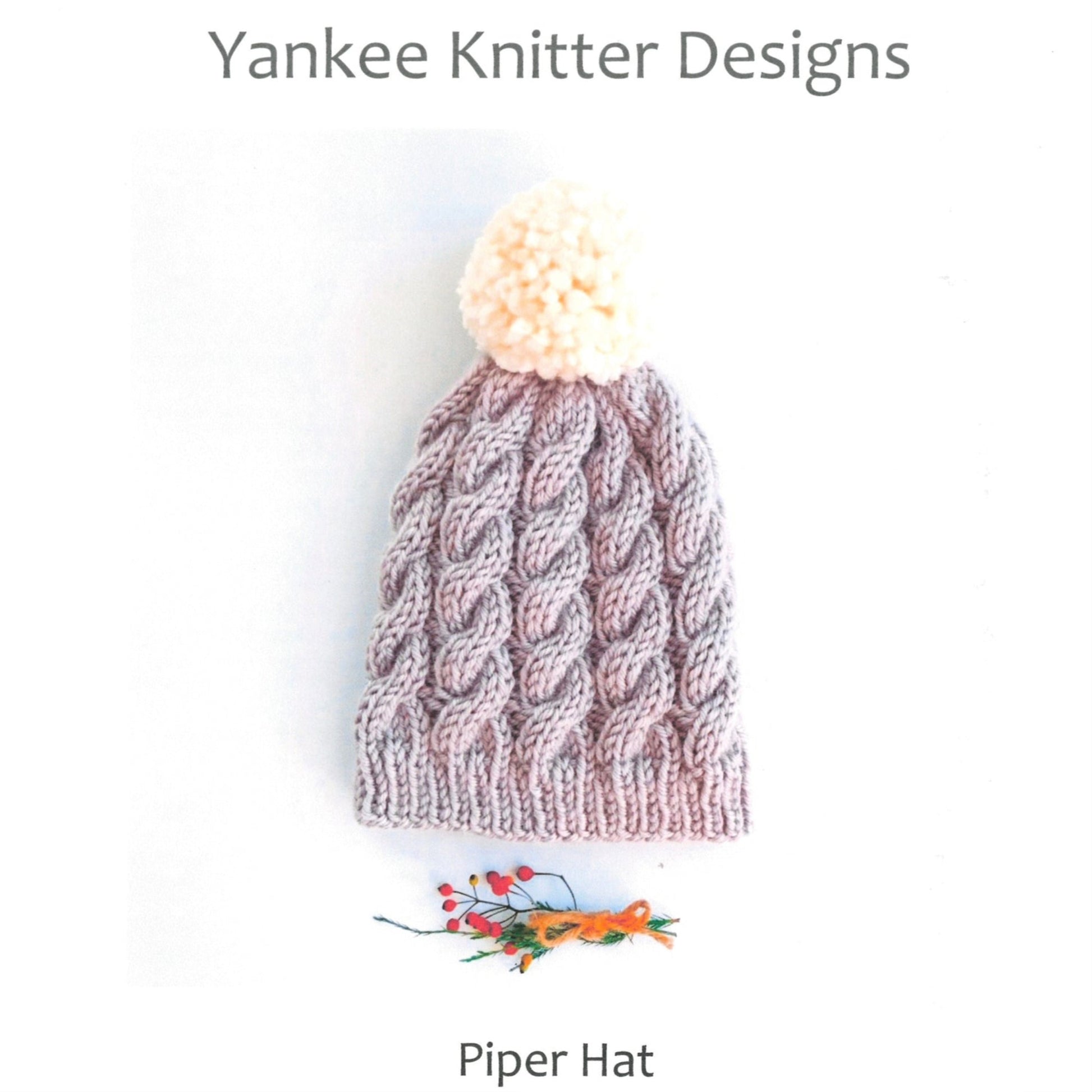 A grey knitted hat with a large white pom-pom on top. The Piper Hat pattern by Yankee Knitter Designs features a cable-knit design and ribbed edges, perfect for those wanting to practice cables. An illustration of flowers is shown below the hat, with "Yankee Knitter Designs" at the top and "Piper Hat" at the bottom.