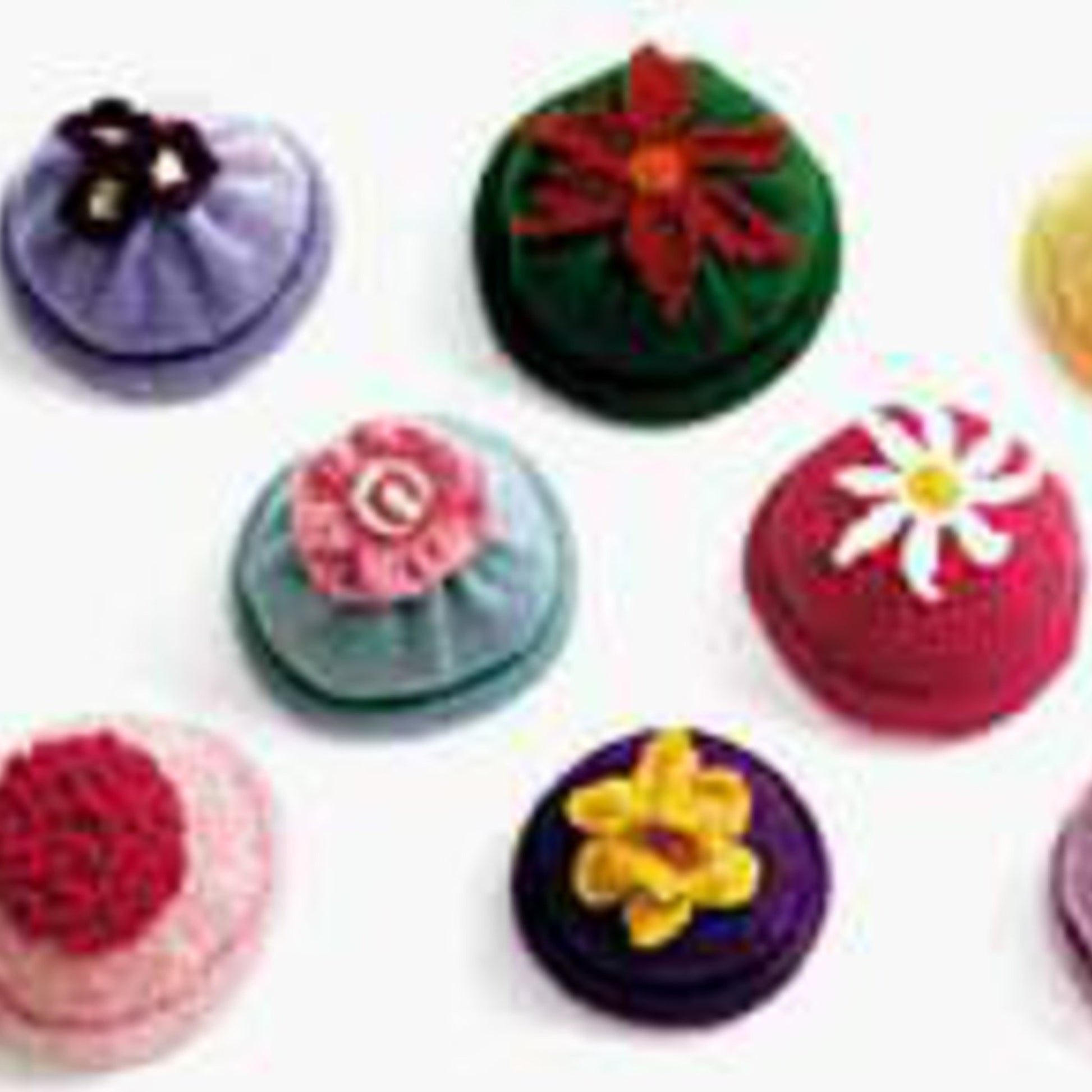 Assorted handmade pincushions are displayed, each uniquely decorated with vibrant fabric flowers and buttons. The collection features various colors and shapes, including round and textured designs with intricate details that evoke the playful charm of How 2 Books' Kid's Flower Caps.