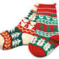 The Nancy Lindberg Christmas Socks Stocking features a pair of socks adorned with charming holiday designs, showcasing festive icons such as snowmen, Christmas trees, gingerbread men, and snowflakes. The vibrant patterns in red, green, white, and yellow make each sock distinct—one predominantly red and green while the other is primarily white and green.