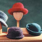 Three felt hats with brim variations, crafted from worsted weight wool by Bryson Distributing, Inc., are displayed on wooden stands against a green backdrop. The center hat is red and placed on a head-shaped stand. The left hat, partially visible, is gray, while the right hat is blue-green and placed on a flat base.
