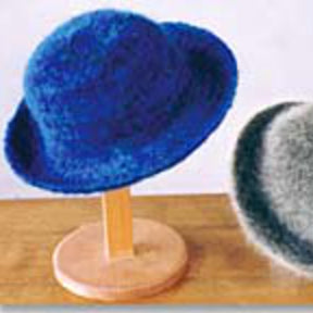 The Felt Hat II - Single Strand by Bryson Distributing, Inc., a blue fuzzy hat made of worsted weight wool, is displayed on a wooden stand on a wooden surface. Another gray and black fuzzy item is partially visible to the right of the blue hat.