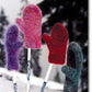 Four vibrant Snow Country Felt Mittens by Bryson Distributing, Inc. (purple, pink, red, and green) are displayed on sticks, standing upright against a snowy backdrop with trees. The mittens are dusted with snow, accentuating the wintry ambiance. These exquisitely crafted accessories complement both women's and men's winter wardrobes perfectly.