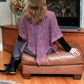 A person with long hair, wearing a light purple Crocheted Cobweb Shawl from Bryson Distributing, Inc., sits on a brown leather ottoman in front of a fireplace. The person is seated with their back to the camera, facing a wood-paneled fireplace with a floral arrangement on a table beside it, embracing the warmth on a cool summer evening.