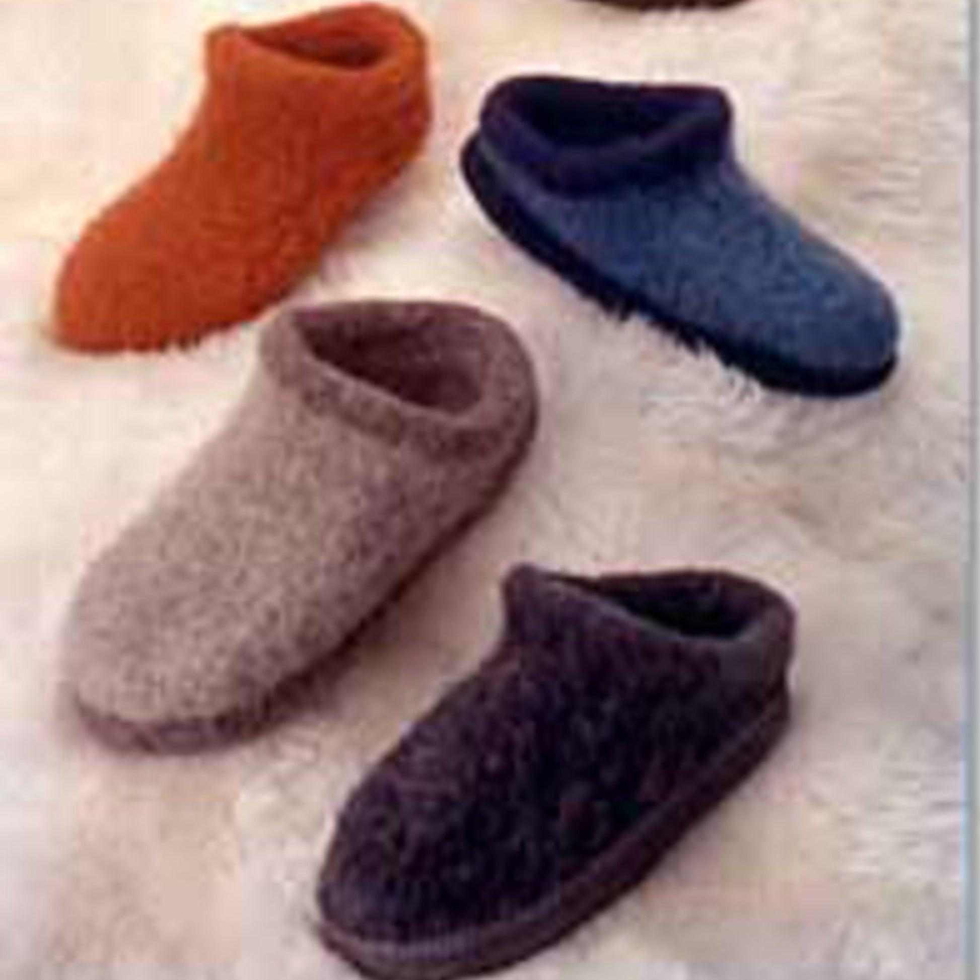 A collection of four Felt Clogs by Bryson Distributing, Inc. arranged on a soft, white surface. The clogs come in various colors: orange, blue, light brown, and dark brown. Each clog features a thick sole and a rounded design, offering a cozy appearance crafted with felted knitting techniques.