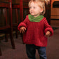 A young child with blonde hair tied in pigtails is wearing a Back Zip Baby Jacket from Bryson Distributing, Inc. They are standing indoors near a row of wooden chairs, smiling, their cozy red jacket with a green collar and blue pants exuding warmth and comfort. The background is softly lit, blending into a warm ambiance.