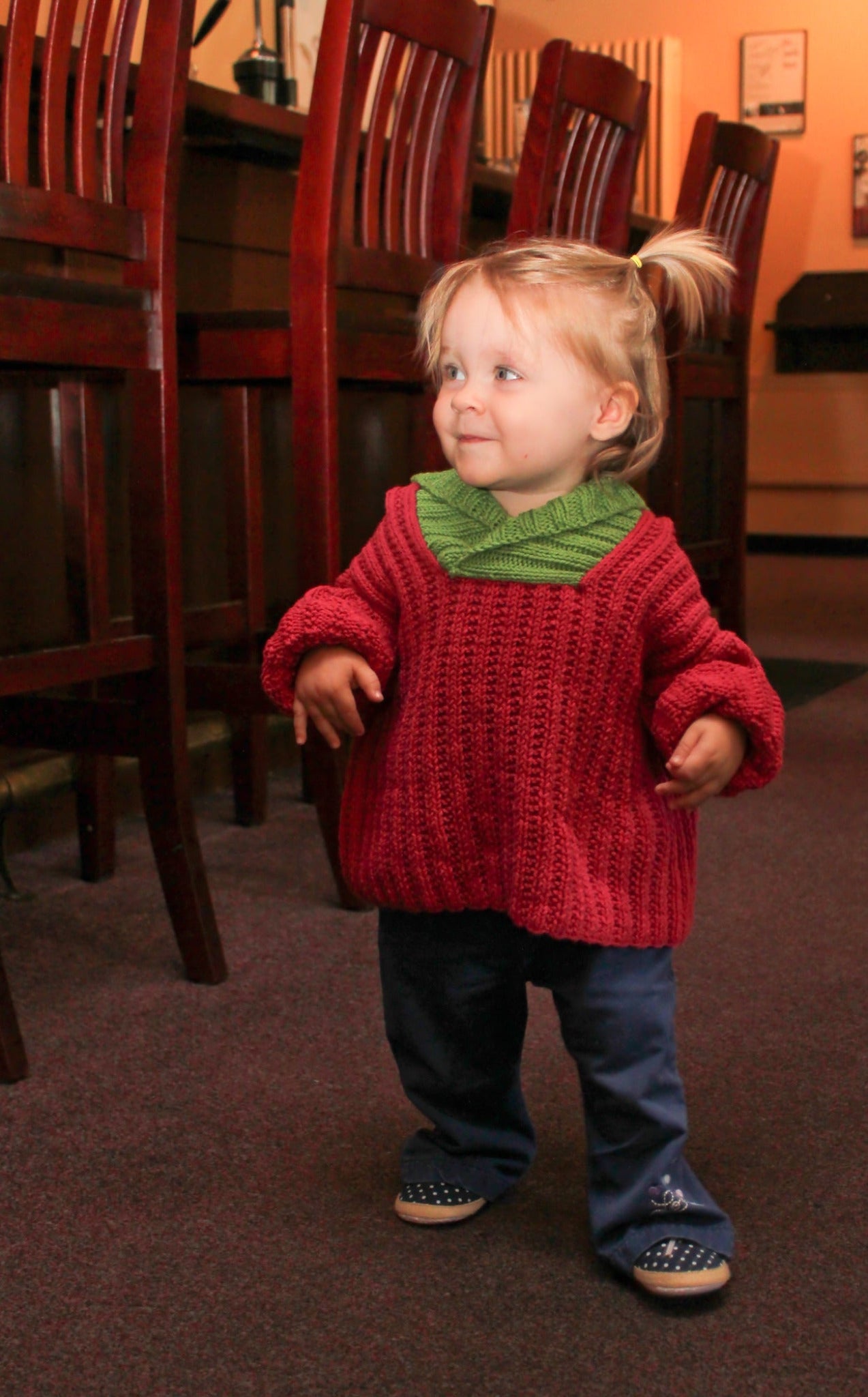 A young child with blonde hair tied in pigtails is wearing a Back Zip Baby Jacket from Bryson Distributing, Inc. They are standing indoors near a row of wooden chairs, smiling, their cozy red jacket with a green collar and blue pants exuding warmth and comfort. The background is softly lit, blending into a warm ambiance.