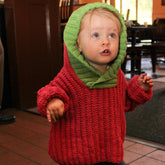 A toddler stands indoors wearing the Back Zip Baby Jacket from Bryson Distributing, Inc. The chunky red jacket with a green hood gives the appearance of a strawberry. The child has light hair and looks up with an expression of curiosity. The background is slightly blurred, showing a tiled floor and furniture.
