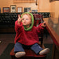 A young child with light hair and a green Back Zip Baby Jacket from Bryson Distributing, Inc. over a red sweater sits on a chair in a cozy room. The child, reaching up with one hand while looking upward, wears the jacket's back zipper open. Framed pictures adorn the walls, and a plant rests on a shelf in the corner.