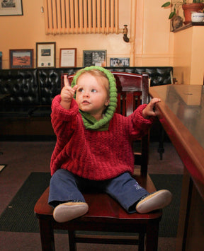 A young child with light hair and a green Back Zip Baby Jacket from Bryson Distributing, Inc. over a red sweater sits on a chair in a cozy room. The child, reaching up with one hand while looking upward, wears the jacket's back zipper open. Framed pictures adorn the walls, and a plant rests on a shelf in the corner.